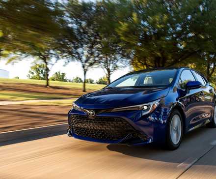 Plan dynamique 3/4 avant d'une Toyota Corolla Hatchback SE 2025 bleue roulant sur une route bordée d'arbres.