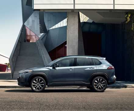Photo de profil latérale statique d'une Toyota Corolla Cross XLE 2025 grise garée à l'extérieur d'un bâtiment moderne.
