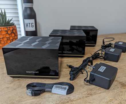 Contenu de la boîte du système Netgear Nighthawk MK93S Tri-Band Mesh Wifi 6E disposé en rangée sur une table.