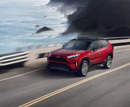 Plan dynamique 3/4 avant d'un Toyota RAV4 Prime XSE 2024 rouge roulant sur une route longeant l'océan.