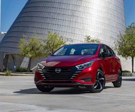 Vue 3/4 avant d'une Nissan Versa 2025 rouge avec un bâtiment moderne en verre en arrière-plan.