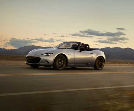 Plan dynamique 3/4 avant d'une Mazda MX-5 2024 argentée roulant sur une route de campagne avec des montagnes en arrière-plan.