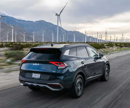 Plan 3/4 arrière dynamique d'un Kia Sportage hybride 2025 bleu roulant sur une route de campagne avec des montagnes et des éoliennes en arrière-plan.
