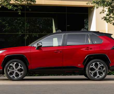 Photo de profil latérale statique d'un Toyota RAV4 PHEV XSE 2025 rouge garé à l'extérieur d'un bâtiment moderne.